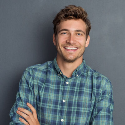 Young handsome man leaning against grey wall with arms crossed. Cheerful man laughing and looking at camera with a big grin. Portrait of a happy young man standing with crossed arms over grey background.