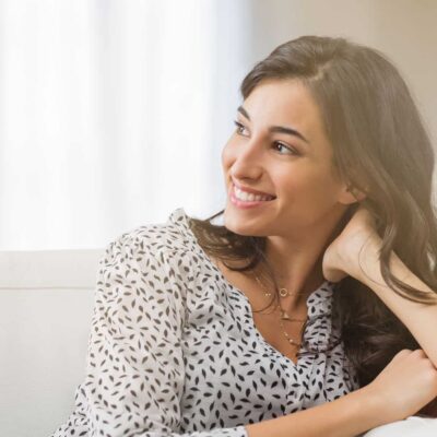 Confident woman relaxing on couch at home. Thoughtful smiling woman sitting on sofa and looking away. Portrait of satisfied girl daydreaming at home.